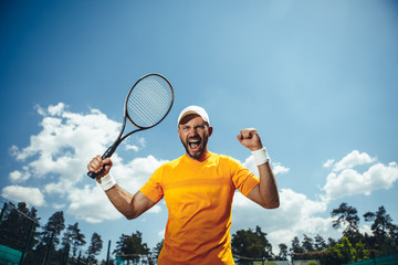 Portrait of cheerful unshaven man gesticulating arm with racket while playing tennis on field outdoor. He shouting and looking at camera