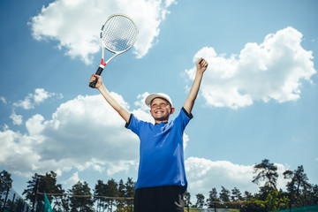 Fototapeta na wymiar Low angle portrait of beaming kid with racket in arm expressing happiness under clear sky. He playing tennis