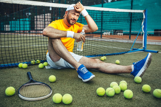 Full length portrait of happy tired bearded male athlete drinking bottle of water while sitting near net outdoor. Green balls and racket locating near him. Satisfied sportsman after training concept