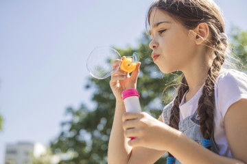 Cute little 10 years old girl in casual outfit playing at park in warm summer day. She blows soap...