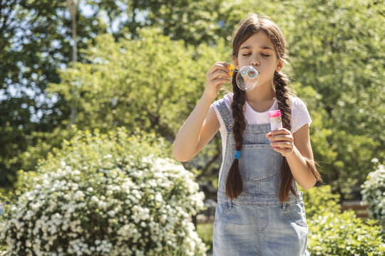 Cute little 10 years old girl in casual outfit playing at park in warm summer day. She blows soap bubbles. Copy space
