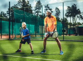 Full length portrait of concentrated bearded male couch playing with focused kid on contemporary court. They keeping rackets in hands