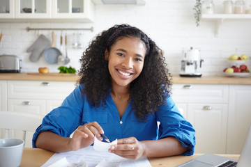 People, paperwork and modern technology concept. Picture of joyful young African woman holding pen and bill while calculating family budget at home, sitting in kitchen with papers and laptop on table - Powered by Adobe