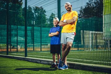 Full length portrait of beaming little boy and happy male wearing sports clothes while situating outdoor. They having rest during training