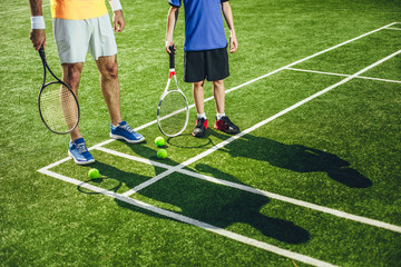 Obraz premium Male trainer and youth athlete holding rackets in arms while standing on green court. Their shadows situating there