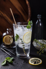 A glass of lemonade with ice with a spoon, an empty bottle and a wooden tray behind and a lemon under a glass bell and a plate with thyme on its side.