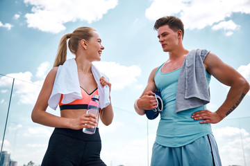 Low angle of cheerful woman and man standing and talking after training in open air. Girl is holding bottle of water while guy is keeping boxing gloves. They are using towels on shoulders and looking