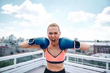 Waist up portrait of angry lady screaming with expression. She is having combat training on terrace of high building in city center. Girl is wearing gloves and holding them near face for showing