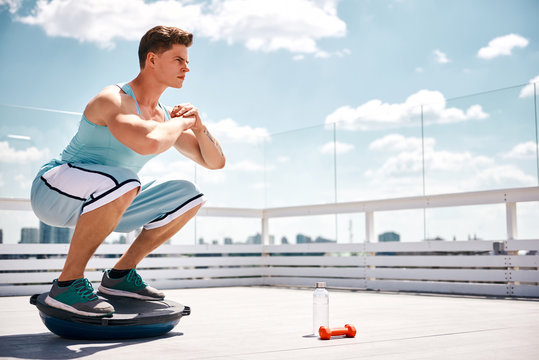 Athlete is doing sit-ups and balancing on BOSU ball. He is exercising on sunny terrace in skies above city. Copy space in right side
