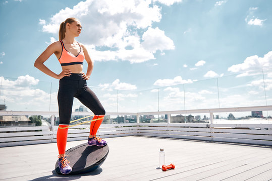 Concentrated fir lady is exercising with dumbbell and resistance band on sunny terrace of skyscraper. She is balancing on BOSU platform and looking forward. Copy space in right side