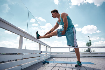 Fit young man is stretching leg while standing and putting it on roof railing. He is getting ready for training outdoors with dumbbells. Male is working out in fresh air concept