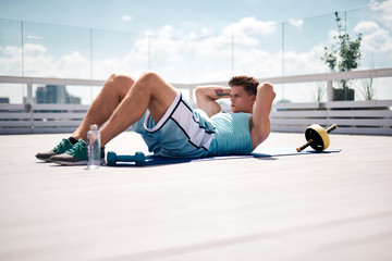 Naklejka premium Fit guy is doing abs crunches on mat in terrace of urban building. He is spending time exercising in open air with sport equipment and water. Work out outdoors on sunny day concept
