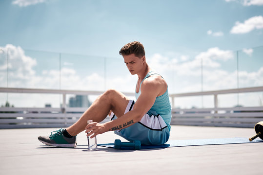 Exhausted athlete is sitting on mat and holding bottle with water. He is going to quench thirst and have short break in training. Male is exercising with dumbbells on roof of skyscraper o sunny warm