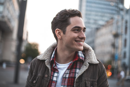 Side View Of Smiling Attractive Male Standing On Street And Looking Sideways. He Is Waiting For His Friend With Joy