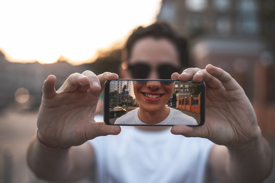 Focus on male hands holding mobile outdoors and making selfie. Young guy is happy and delighted