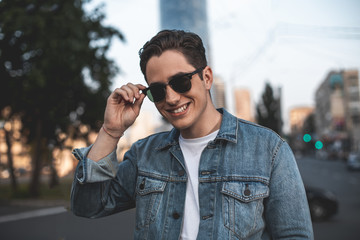Waist up portrait of smiling young attractive man standing on street. He is touching rim of sunglasses with delight