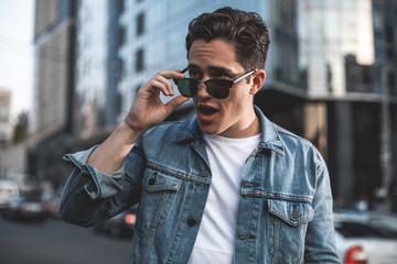 Waist up portrait of astonished male standing outside and putting off sunglasses. He is trying to...