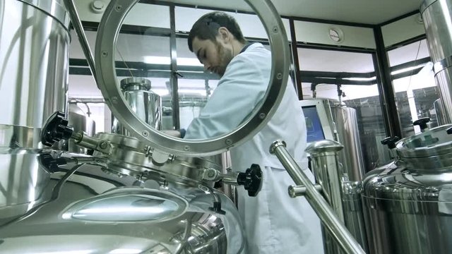 Professional worker in lab coat using steel tool for stirring beer in tun during brewing process