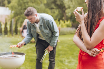 Fototapeta premium Happy woman eating bread while her friend grilling shashliks