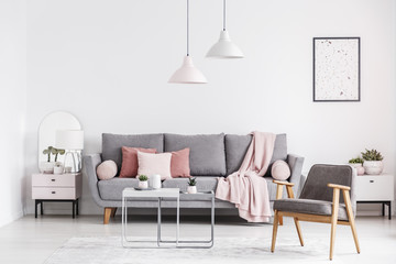 Wooden armchair and poster in white living room interior with pink blanket on grey couch. Real photo