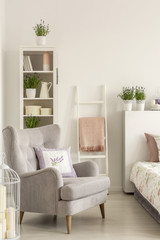 Grey armchair with a pillow, ladder with a blanket and shelf with decorations in a bedroom interior. Real photo