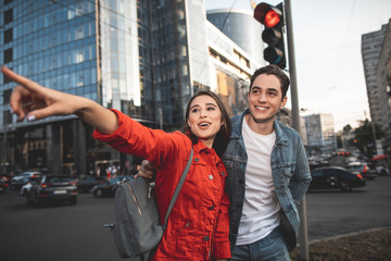 Waist up portrait of delighted girl pointing finger on street. Happy boyfriend is standing near her looking with content