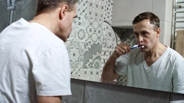 Medium shot of middle-aged man in white t-shirt looking in the mirror while brushing teeth after waking up in the morning