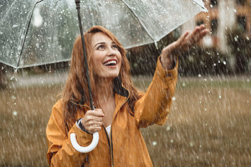 Waist up portrait of cheerful girl holding umbrella and catching rain drops with smile. She is...