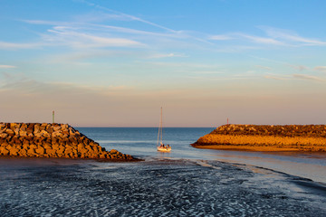 a sail boat leaving the harbour at low tide, as the sun is setting, radiating an orange glow