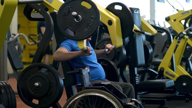 PAN of disabled sportsman exercising on chest press machine in gym; wheelchair standing beside him