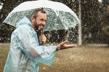 Side view of laughing man holding umbrella and catching water drops outdoors. He is standing on grass and smiling with excitement