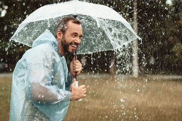Side view of smiling happy man standing on grass and holding parasol. He is feeling blissful about wet rainy weather. Copy space in right side