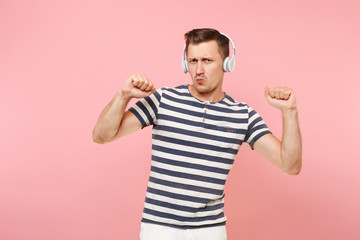 Portrait of smiling young man wearing striped t-shirt listening to music with white wireless headphones, enjoy isolated on trending pastel pink background. People sincere emotions lifestyle concept.