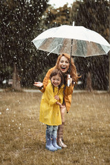 Full length of joyful child standing near mother in raincoat. Content woman is holding umbrella and...