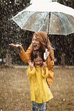 Full length of happy mother standing under umbrella with small girl. She is stretching hand trying to catch water drops with closed eyes in delight