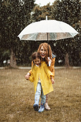 Full length of little child standing by her smiling mother and hiding from rain under umbrella. They are happy and excited