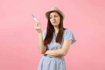 Sad unhappy woman in blue dress, hat hold in hand, looking at pregnancy test isolated on pink...