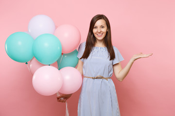 Portrait of fascinating young happy woman in blue dress holding colorful air balloons pointing hand aside on copy space isolated on bright trending pink background. Birthday holiday party concept.