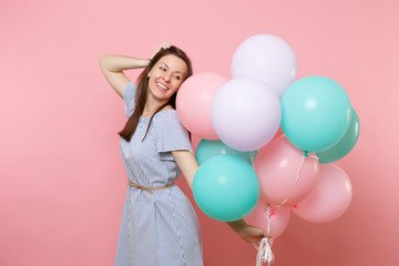 Portrait of tender smiling young woman in blue dress holding colorful air balloons keeping hand near head isolated on bright pink background. Birthday holiday party, people sincere emotions concept.