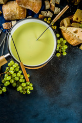 Gourmet Swiss fondue in traditional fondue pot, with forks, various cheeses, olives, bread and grape, dark blue background copy space