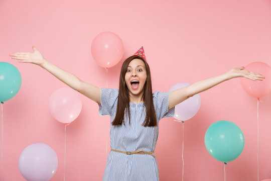 Portrait of amazed young happy woman in birthday hat blue dress screming spreading hands on pastel pink background with colorful air baloons. Birthday holiday party, people sincere emotions concept.