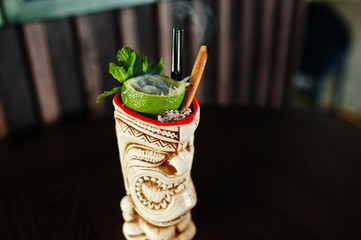 Alcoholic cocktail with mint and lime with fire in originally voodoo earthen glass on bar table.