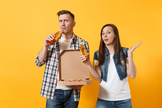 Young concerned couple woman man sport fans cheer up support team drinking beer hold italian pizza in cardboard flatbox spreading hands isolated on yellow background. Sport family leisure lifestyle.