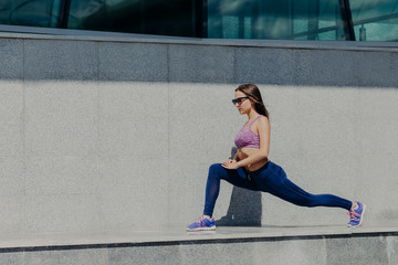 Fototapeta na wymiar Healthy lifestyle and sport concept. Young fit female runner does stretching exercises after running in morning, shows her flexible perfect body, wears comfortable sneakers, shades and purple top