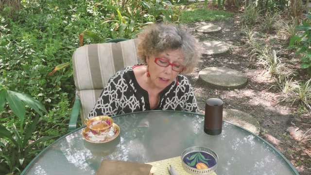 An active senior woman drinking tea in her garden looks up at the sky to check the weather before consulting her smart home device. Variation with no book and brown smart home device