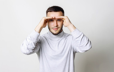 Studio portrait of handsome positive male keeping his hands over his eyes to protect from the sunrays. Bearded man looking at the camera keep his hands on forehead on studio wall background. People