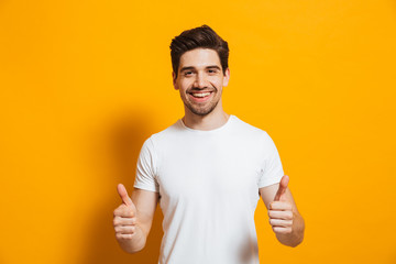 Portrait of cheerful man in basic clothing smiling and showing thumbs up at camera, isolated over...