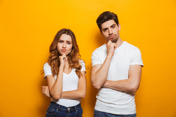 Photo of resented man and woman in casual clothes standing together and touching chin with irritated look, isolated over yellow background