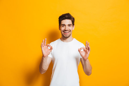 Portrait of handsome excited man in basic clothing smiling and showing ok sign at camera, isolated over yellow background