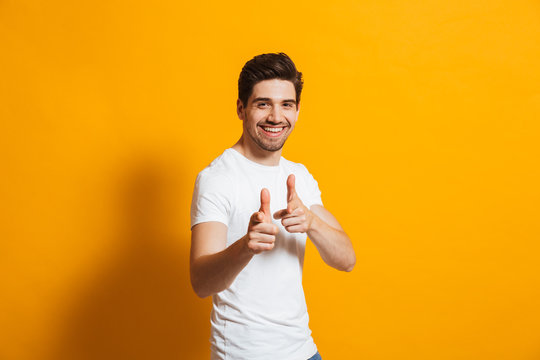 Image of pleased guy in white t-shirt laughing and pointing finger at camera, isolated over yellow background